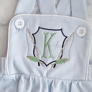 May include: Light blue baby overalls with a white trim and a white button. The overalls have an embroidered design of a shield with a letter K, two fishing rods, and two fish. The text "FISHING CREST" is at the top of the image. The text "FONT NOT INCLUDED" and "STITCHED BY @THEOLLIEPOPSHOP" is at the bottom of the image.