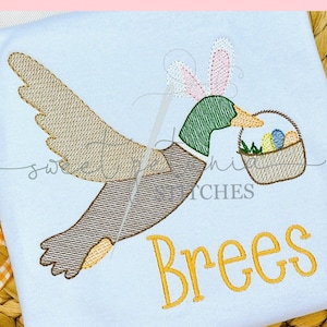 May include: White t-shirt featuring an embroidered Easter duck wearing bunny ears and carrying a basket of Easter eggs. The name "Brees" is embroidered in yellow. Text at the top reads "EASTER DUCK".