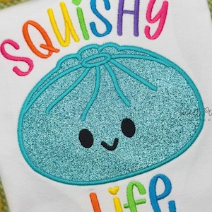 Peut inclure: Un t-shirt blanc avec une broderie représentant un personnage de boulette bleu sarcelle à paillettes avec un visage souriant. Au-dessus, le mot SQUISHY est brodé en lettres multicolores, et en dessous, le mot LIFE est brodé en lettres multicolores.