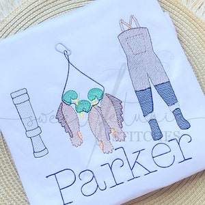 Puede incluir: Camiseta blanca con diseño bordado de temática pesquera. Presenta una caña de pescar, botas de agua y una ristra de peces. El nombre "Parker" está bordado en azul. El diseño incluye detalles verdes, marrones y azules.