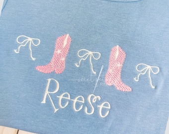 Könnte beinhalten: Ein hellblaues T-Shirt mit rosa gestickten Cowboystiefeln und Schleifen. Der Name "Reese" ist in Weiß unter den Stiefeln und Schleifen gestickt.