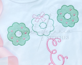 Op de afbeelding: Een wit shirt met drie groene geborduurde kransen en een roze geborduurde letter E. De kransen hebben roze stippen en één krans heeft een roze strik.