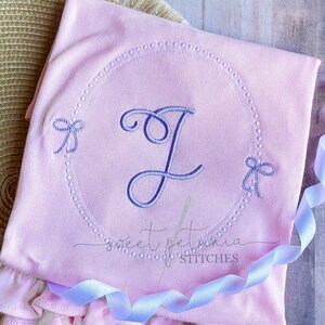 May include: A pink baby onesie with a pearl-like circle design and a cursive letter J in the center. The onesie has two small bows on either side of the circle. The text "sweet petunia stitches" is at the bottom of the onesie.
