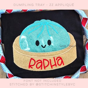 Puede incluir: Una camiseta negra con una aplicación bordada de una bola de masa brillante de color turquesa con una cara sonriente en una cesta de vapor de bambú beige. El nombre Rapha está cosido con hilo rojo en la parte delantera de la cesta. El diseño está centrado.