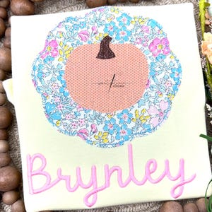 Könnte beinhalten: Hellgelbes T-Shirt mit einem rosa Kürbis-Applikation. Der Kürbis hat einen braunen Stiel und ist von einem Blumenmuster in Blau, Rosa und Gelb umgeben. Der Name "Brynley" ist darunter in rosa Schrift gestickt.