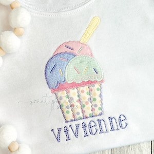May include: A white t-shirt with a colorful embroidered cupcake design and the name "Vivienne" stitched in purple thread.