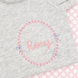 May include: A light gray baby onesie with the name "Rory" embroidered in pink script within a pink dotted circle. Small blue bows accent the circle. The words "sweet peonia stitches" are below the name.