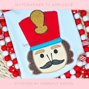 May include: White shirt with a colorful appliqué of a nutcracker soldier. The nutcracker has a red hat with a gold accent, a black mustache, and a tan face. The text "NUTCRACKER ZZ APPLIQUE" and "STITCHED BY KIMBERLY GWYNN" are visible.