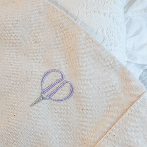 May include: A white canvas tote bag with a purple embroidered heart-shaped scissors design.