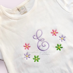 May include: White t-shirt with a purple embroidered letter E and pink, green, and yellow embroidered flowers. The text "sweet petunia stitches" is embroidered below the letter E.