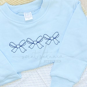 Puede incluir: Sudadera azul claro con tres lazos azules bordados y el texto "sweet petunia STITCHES".