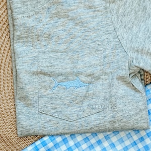 May include: A gray t-shirt with a blue embroidered swordfish design on the pocket. The t-shirt is folded and sitting on a checkered blue and white surface.