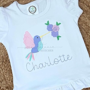 May include: White short-sleeved shirt with a hummingbird and flowers embroidered on the front. The name "Charlotte" is embroidered below the hummingbird.