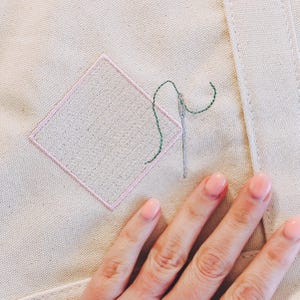 May include: A close-up of a hand holding a needle and thread over a pink diamond-shaped patch of fabric on a beige canvas background. The thread is green and the needle is silver.