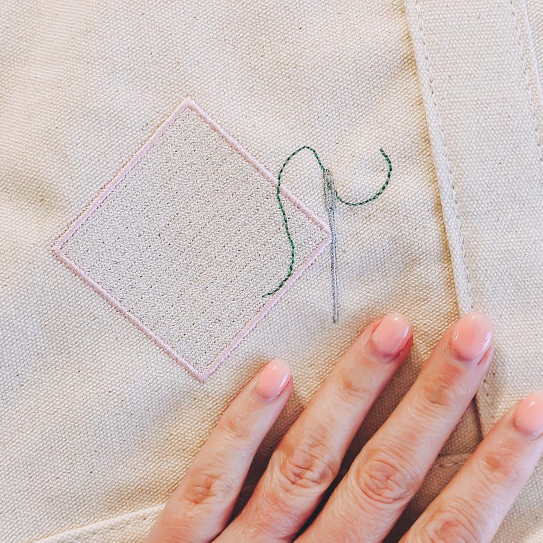 May include: A close-up of a hand holding a needle and thread over a pink diamond-shaped patch of fabric on a beige canvas background. The thread is green and the needle is silver.