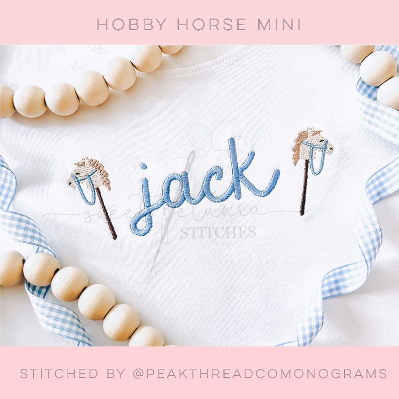 May include: A white onesie with blue embroidery of the name "Jack" and two hobby horses on either side. The onesie is decorated with a blue and white gingham ribbon and wooden beads.