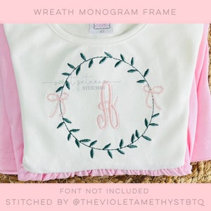May include: A white long-sleeved shirt with a pink ruffle hem. The shirt features a green embroidered wreath with a pink bow and a pink monogram letter. The text "WREATH MONOGRAM FRAME" is at the top.