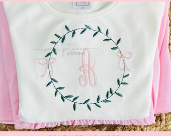 May include: A white long-sleeved shirt with a pink ruffle hem. The shirt features a green embroidered wreath with a pink bow and a pink monogram letter. The text "WREATH MONOGRAM FRAME" is at the top.