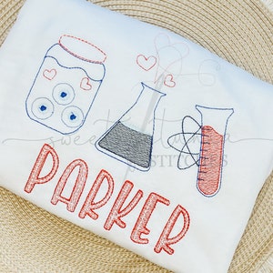 May include: A white t-shirt with red embroidered text that says "Parker". The design also includes a jar with blue and white eyes, a beaker with a blue liquid, and a graduated cylinder with red liquid. There are also red hearts and a red and blue atom symbol.