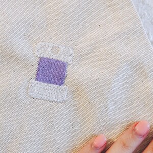 May include: A white embroidered spool of thread with a purple thread wound around it.