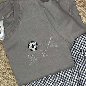 May include: A gray t-shirt with a white soccer ball and the name "Jack" embroidered on it. The shirt is laying on top of a black and white checkered fabric.