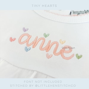 Op de afbeelding: Wit kledingstuk met de naam "Annie" geborduurd in een zachte perzikkleur. Kleine pastelkleurige hartjes in roze, groen, blauw en geel omringen de naam. De tekst "Tiny Hearts" staat bovenaan en "Sweet Petunia Stitches" staat in de afbeelding.