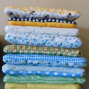 May include: A pile of rectangular fabric items in various patterns and colours. Designs include bees, polka dots, gingham, and floral prints. The colours are yellow, blue, green, and white. The items appear to be pillows or cushions.