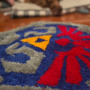 Op de afbeelding: Een grijs, blauw, rood en geel gebreid tapijt met een gestileerd ontwerp van een gevleugeld symbool. Het tapijt is gemaakt van zacht, pluche garen.