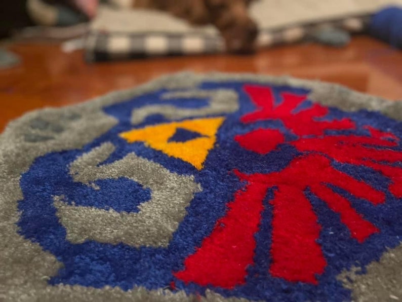 Op de afbeelding: Een grijs, blauw, rood en geel gebreid tapijt met een gestileerd ontwerp van een gevleugeld symbool. Het tapijt is gemaakt van zacht, pluche garen.