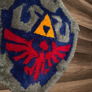 Op de afbeelding: Een blauw, rood en geel gehaakt tapijt dat het Triforce-symbool uit de videogame-serie The Legend of Zelda afbeeldt. Het tapijt is schildvormig en heeft een grijze rand.