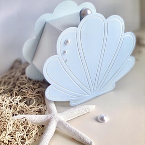 Könnte beinhalten: Hellblaue Muschelschachtel mit Perlenakzent, weißem Seestern und Perle. Die Schachtel hat einen gewellten Rand und ein detailliertes Muscheldesign. Ein Fischernetz im Hintergrund erzeugt ein Strandthema.