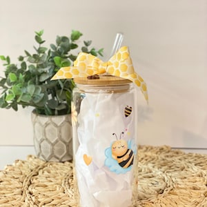 Puede incluir: Un tarro de vidrio transparente con tapa de bambú y una cinta amarilla de panal de abeja atada alrededor de la tapa. El tarro tiene un diseño de abeja de dibujos animados lindo con un esquema de color azul y amarillo. El tarro está lleno de papel de seda blanco.