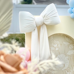 May include: A large white bow made of fabric is tied to a light yellow backdrop with a floral pattern. The bow is tied in a knot and the tails are hanging down. There are pink and brown balloons in the background.