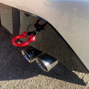 May include: A red tow hook with a black metal ring attached to a silver car. The car has two chrome exhaust pipes.