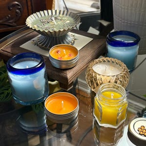 Könnte beinhalten: Eine Auswahl an Duftkerzen in verschiedenen Behältern. Enthält Kerzen in blauem Glas, Metalltöpfen und einem dekorativen goldfarbenen Glashalter. Die Kerzen sind in Blau-, Orange- und Gelbtönen gehalten.