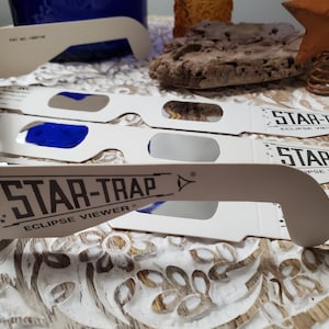 May include: A set of four white cardboard eclipse viewers with the text "STAR-TRAP ECLIPSE VIEWER" printed on them. The viewers are designed to be held up to the eyes to safely view a solar eclipse.