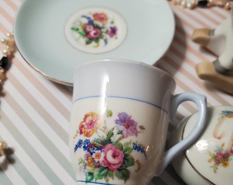 May include: A blue and white teacup and saucer set with a floral design. The cup has a white background with a blue band and a floral design in shades of pink, yellow, and green. The saucer is light blue with a white center and a floral design in the center.