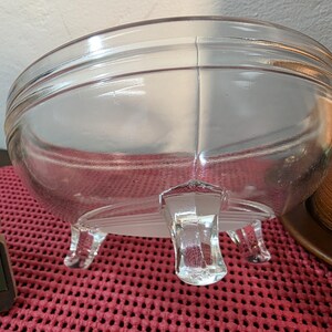 May include: A clear, glass bowl with a rounded shape, resting on three small, clear feet. The bowl has a slight lip and a seam running down the center. The bowl is sitting on a red, textured surface.