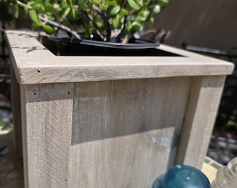 May include: A weathered, square wooden planter box with a live green plant inside. The box has a light gray finish and is constructed with visible wood grain. A blue glass insulator sits in front of the planter.