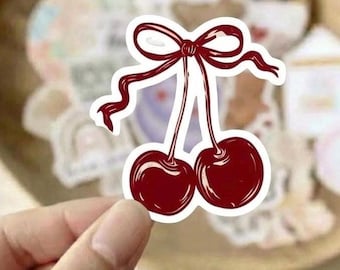 Puede incluir: Una pegatina troquelada que muestra dos cerezas de color rojo oscuro conectadas por tallos a un lazo decorativo de cinta roja. La ilustración utiliza contornos marcados y reflejos blancos para crear un aspecto brillante sobre un borde blanco.
