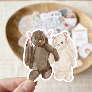 May include: A die-cut sticker featuring a watercolor illustration of two plush bunnies holding hands. The left bunny is dark brown and the right bunny is cream-colored. Both bunnies have long ears and are wearing small, light pink ribbon bows.