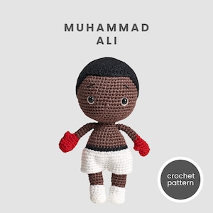 Puede incluir: Patrón de crochet para una muñeca amigurumi marrón y negra de Muhammad Ali con guantes de boxeo rojos y pantalones cortos blancos. La muñeca tiene un afro negro y ojos marrones. El texto "crochet pattern" está en un círculo en la parte inferior derecha de la imagen.