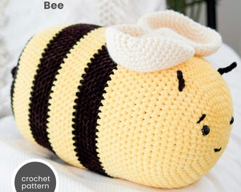 Puede incluir: Una almohada con forma de abeja tejida a crochet con rayas negras y amarillas. La abeja tiene un ala blanca tejida a crochet y ojos negros. La almohada está sobre una superficie blanca. El texto "Snuggle-Sized Bee" y "crochet pattern" es visible en la imagen.