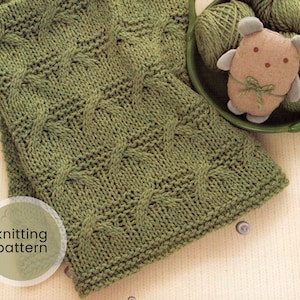 Può includere: Una copertina per neonati di colore verde a maglia con un motivo a treccia. La copertina è appoggiata su una superficie marrone chiaro con un piccolo peluche.