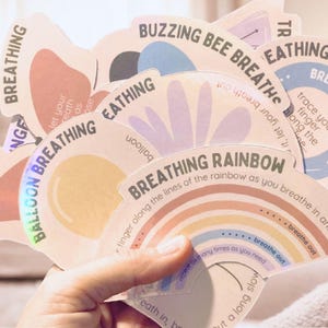 May include: A hand holds a fan of illustrated cards with breathing exercises. The cards feature colourful designs and text like "Butterfly Breathing" and "Breathing Rainbow." The cards are designed to help with mindfulness and relaxation.