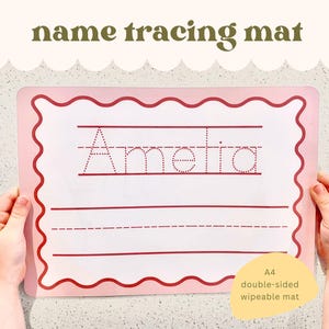 May include: A pink A4 name tracing mat with a wavy border. The name "Amelia" is printed in dotted lines for tracing, with lines below. The mat is double-sided and wipeable, with the text "name tracing mat" at the top.