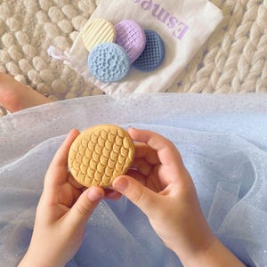 May include: A child's hands hold a yellow, textured, round object with a scale-like pattern. A small bag with the name "Esme" is visible, along with several other textured, round objects in various colours. The objects appear to be toys.