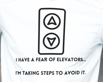 Puede incluir: Camiseta blanca con un gráfico negro de un botón de ascensor con flechas arriba y abajo. El texto debajo del gráfico dice "Tengo miedo a los ascensores... Estoy tomando medidas para evitarlo."