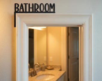 Może przedstawiać: Czarny metalowy znak "BATHROOM" zamontowany nad białą ramą drzwi. Znak jest w kursywnym kroju pisma i jest przymocowany do ramy drzwi za pomocą wspornika kątowego. Łazienka jest widoczna przez otwór drzwiowy.