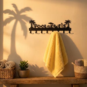 May include: A black metal towel rack with the words "POOL TOWELS" cut out, decorated with palm trees and beach icons. A yellow towel hangs from the rack. Rolled towels and a small plant sit on a wooden shelf below.